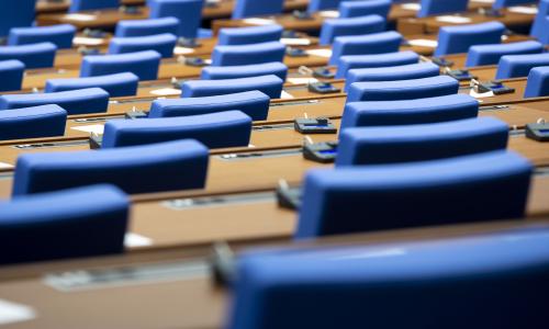 A imagem mostra uma sala de conferências com fileiras de assentos azuis e mesas de madeira